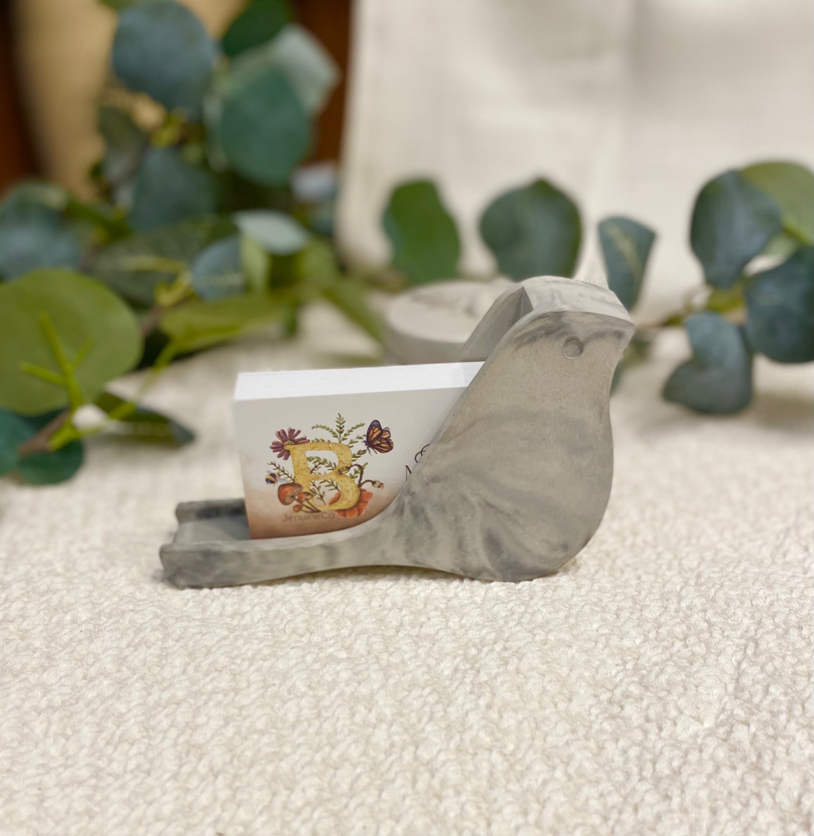 Decorative bird-shaped card holder with a card featuring a floral design on a textured surface with greenery.