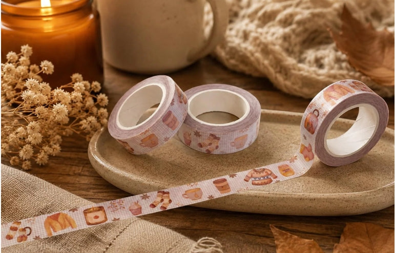 Three rolls of patterned washi tape on a ceramic tray, styled with dried flowers, a candle, and a knitted blanket in a warm autumn setting.